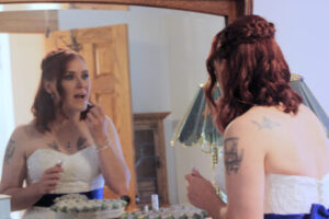 A woman in a strapless white wedding dress applies lip gloss while looking into a mirror.