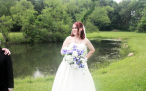 A bride in a white wedding dress holding a bouquet of white and blue flowers in front of a pond.