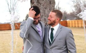 Two men in grey suits smiling at each other during an outdoor wedding ceremony.