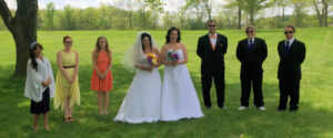 Two brides in white wedding dresses with three bridesmaids and three groomsmen standing in a grassy field.
