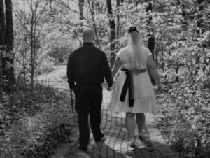 A bride in a short white dress and a groom in a dark shirt walking hand-in-hand along a wooded path.