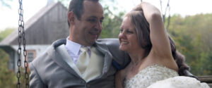 A bride in a beaded white gown and a groom in a grey suit sitting together on a porch swing.