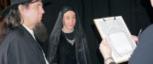 A person in a black veil stands between a man in a black hat and someone holding a decorated document on a clipboard.
