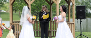An officiant stands between two brides in wedding dresses inside a wooden gazebo during an outdoor ceremony.