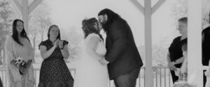 A bride and groom kiss under a gazebo while wedding guests look on.