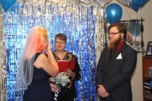 An officiant leads a wedding ceremony for a bride and groom in front of a blue tinsel backdrop.