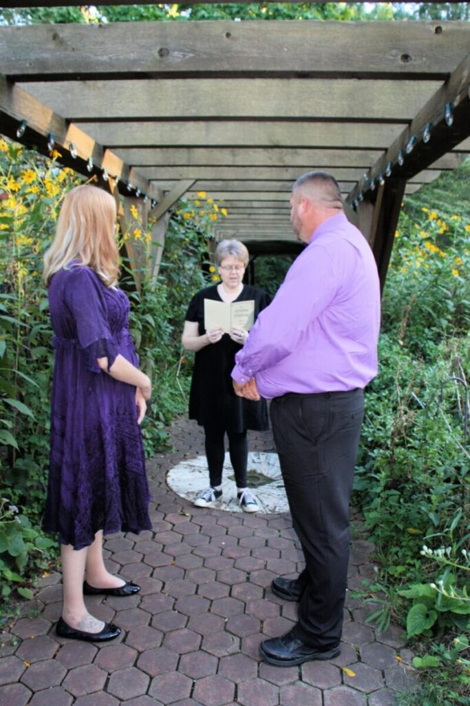 An officiant reading from a book during an outdoor wedding ceremony between a woman in purple and a man in a purple shirt.