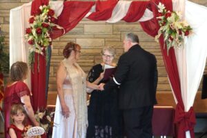 A bride and groom hold hands while an officiant leads their wedding ceremony under a decorated archway.