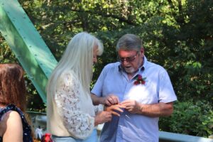 A woman with long white hair placing a ring on a man’s finger during an outdoor ceremony.