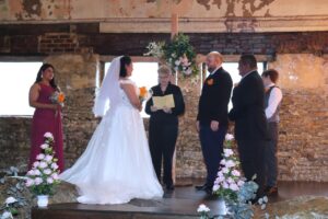 A bride and groom standing at an altar with an officiant and wedding party in an industrial room with exposed brick walls.