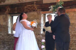 A bride laughing while standing with a groom and an officiant during a wedding ceremony in a rustic room.