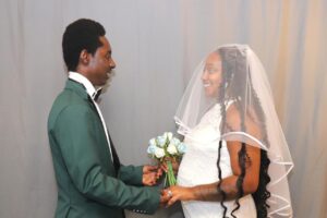 A groom in a green suit and a bride in a white wedding dress and veil hold hands and look at each other.