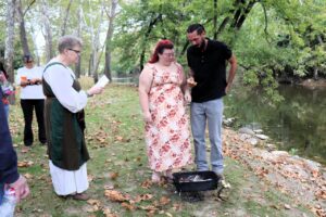 A couple stands before a small portable grill by a river while an officiant reads from a paper.