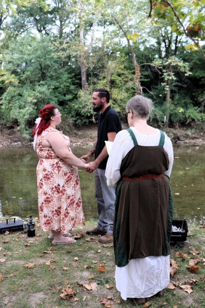 A couple holding hands during an outdoor wedding ceremony officiated by a person in a brown and green gown.
