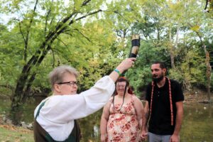 A person in medieval-style clothing holds a drinking horn above a couple standing near a river.