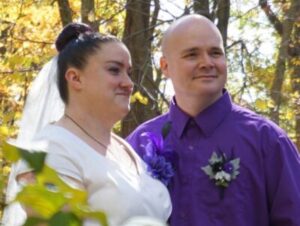 A bride in a white dress and veil stands next to a groom in a purple button-down shirt outdoors.