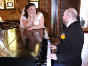 A bride in a wedding dress leans on a piano while the groom plays it.