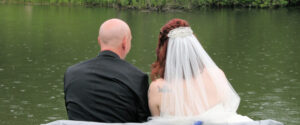 A bride and groom in wedding attire sit side-by-side facing a lake in the rain.