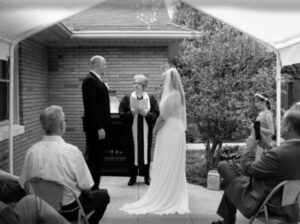 A couple stands before an officiant during an outdoor wedding ceremony, viewed from behind by guests.