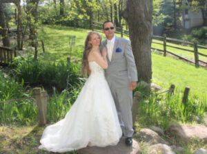 A bride in a white wedding gown and a groom in a grey suit posing together in a garden.