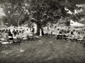 A couple stands beneath a large tree during an outdoor wedding ceremony with guests seated in rows.