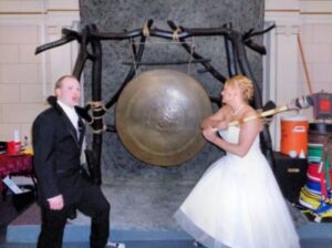 A bride in a wedding dress holds a mallet to strike a large gong, while a man in a tuxedo stands next to her.
