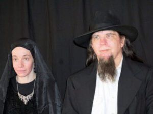 A woman wearing a black veil and necklace next to a man with a goatee wearing a black hat and suit.