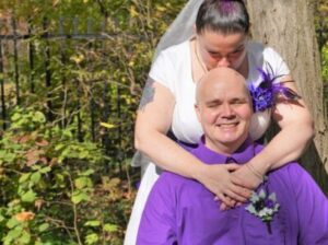 A bride in a white dress stands behind and kisses the head of a groom wearing a purple shirt.