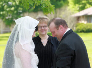 A bride and groom smile at each other during a wedding ceremony with an officiant standing between them.