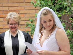 A bride in a veil reads a paper while an officiant smiles beside her.