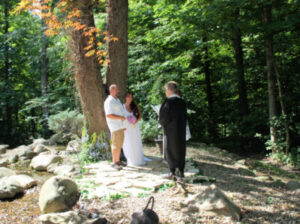 A couple standing in a wooded area during an outdoor wedding ceremony, facing an officiant.
