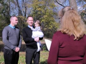Two men standing outdoors with a small child, facing an officiant.