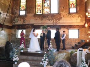 A bride and groom exchange wedding vows on a wooden stage in front of an officiant and two witnesses.