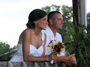 A bride in a white gown and a groom in a white shirt leaning on a wooden railing, looking in the same direction.
