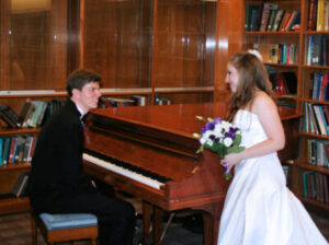 A man in a tuxedo playing a grand piano for a woman in a white wedding dress holding a bouquet in a library.