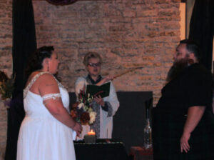 A bride and groom stand before an officiant holding a book and a wand during a wedding ceremony.