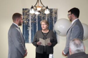 An officiant reads from a document during a wedding ceremony with two grooms in grey suits standing before her.